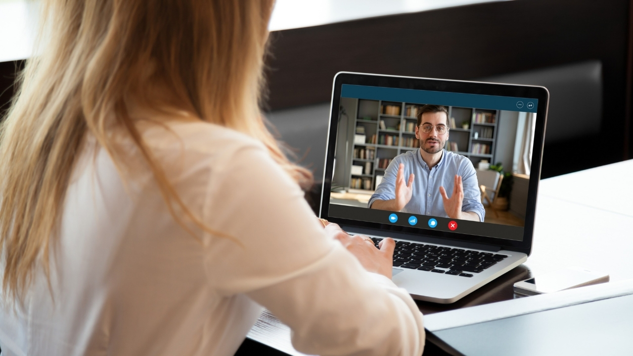 Eine Frau sitzt vor einem Bildschirm, auf dem eine Videokonferenz läuft, in der ein Mann spricht